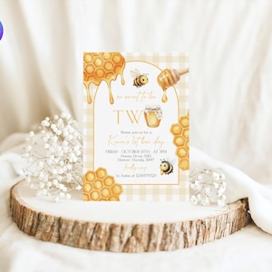 May include: A honey-themed birthday invitation with the text "So sweet to be TWO." The design includes illustrations of honeycombs, bees, and a honey dipper. The invitation is on a light yellow checkered background.
