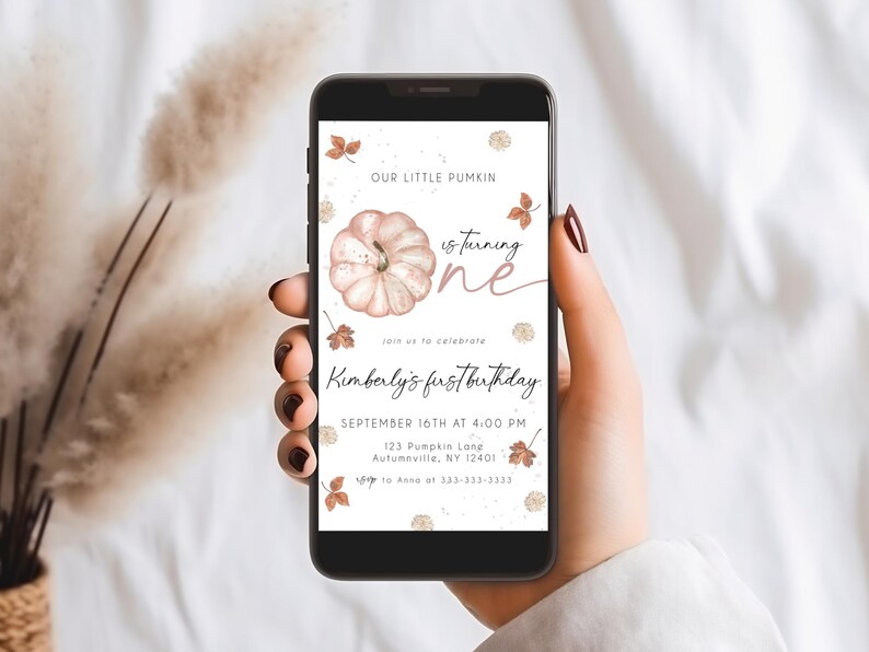 May include: A smartphone displays a digital invitation for a first birthday. The design features a watercolor pumpkin, fall leaves, and the text "Our Little Pumkin is turning one." The invitation includes the name "Kimberly's first birthday" and event details.
