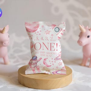 May include: A pink and white party favor bag with an astronaut and space-themed graphics. The bag reads "5, 4, 3, 2... ONE! 1st trip around the sun!" Two pink unicorn figurines flank the bag on a wooden stand.