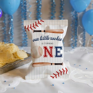 May include: A white chip bag with baseball-themed graphics and text that reads "our little rookie is turning ONE." The bag features baseball bats, a baseball, and red and blue accents. Blue balloons and chips are in the background.