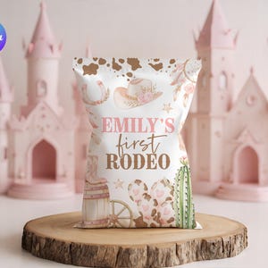 May include: A white party favor bag with a cowgirl theme, featuring a cowboy hat, horseshoe, and cactus illustrations. The text "Emily's First Rodeo" is displayed in pink and brown. The bag is set on a wooden surface.