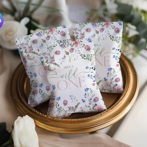 May include: Three white gift bags with floral patterns of pink, purple, and blue flowers, arranged on a gold-rimmed plate. The bags have the text "She's a Wild One" in pink and green script.