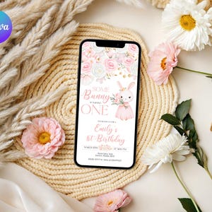 Może przedstawiać: Smartfon wyświetla zaproszenie urodzinowe z ilustracją królika i tekstem "Some Bunny is Turning One". Zaproszenie ma różowe i białe kwiatowe akcenty. Telefon spoczywa na plecionej macie, otoczonej kwiatami i elementami dekoracyjnymi.
