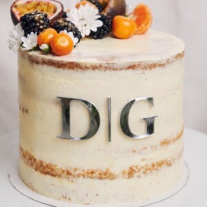 May include: A semi-naked cake with white frosting and the silver letters DIG attached to the front. The top is decorated with fresh blackberries, passion fruit halves, orange physalis, mandarin segments, and small white flowers.