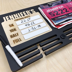 May include: Black wooden race bib and personal best display board. The board has slots for race bibs and a section for recording times. The text "JENNIFER'S PERSONAL BESTS" is displayed, along with times for 5K, 10K, Half, and Full races. A black marker is included.