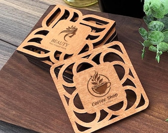 May include: A stack of square wooden coasters with intricate laser-cut geometric borders. The top coaster features a coffee cup graphic and the text Coffee Shop your slogan. Another coaster shows a beauty icon and the text BEAUTY your slogan.