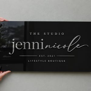 May include: A rectangular black acrylic sign featuring silver 3D lettering that reads THE STUDIO jenninicole EST. 2021 LIFESTYLE BOUTIQUE. The sign is mounted with four black circular standoffs at each corner, creating a sleek, professional display.