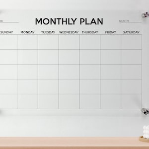 May include: A clear acrylic wall-mounted monthly calendar board featuring black printed text that reads MONTHLY PLAN, YEAR, MONTH, and the days of the week from Sunday to Saturday. The grid is secured to the wall with four silver metal standoffs.
