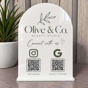 Peut inclure: Un panneau en acrylique blanc en forme d'arche portant le texte Olive & Co. BEAUTY STUDIO, Connect with us, Instagram et Leave a Review. Le panneau comprend un dessin au trait botanique, une icône Instagram, une icône G et deux QR codes.