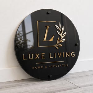 Peut inclure: Une enseigne circulaire en acrylique noir avec des lettres et un logo en relief de couleur dorée. Le panneau affiche le texte LUXE LIVING et HOME & LIFESTYLE, avec une grande lettre L dans un cadre carré à côté d'un motif de branche dorée.