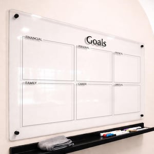 May include: A clear acrylic wall-mounted dry erase board titled Goals with six rectangular sections labeled Financial, Personal, Physical, Family, Career, and Spiritual. It features black mounting hardware and a black shelf holding markers and a cloth.