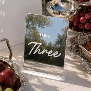 May include: A rectangular, mirrored table sign with a white base. The sign features the word TABLE in a simple sans-serif font above the word Three in a white, cursive script. The reflective surface shows a clear outdoor scene with trees.