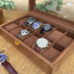 Peut inclure: Une boîte à montres en bois avec un couvercle en verre et huit compartiments pour ranger les montres. La boîte est en bois foncé et est doublée de velours brun. Il y a cinq montres dans la boîte, toutes de styles et de designs différents.