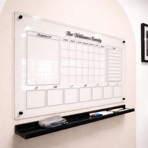 May include: A clear acrylic wall-mounted calendar board with black text reading The Williams Family, featuring a weekly planner, a shopping list section, and a to-do list. It includes a black shelf below holding markers and a cleaning cloth.