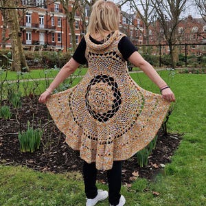 May include: A handcrafted crochet vest in shades of tan, orange, and blue. The vest has a hooded design and a circular pattern on the back. The person is wearing a black top, black trousers, and white trainers.