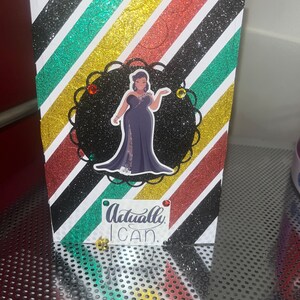 May include: A greeting card with a black, gold, red, and green striped background. The card features a black and white illustration of a woman in a black dress with the text "Actually, I can." in black and white.