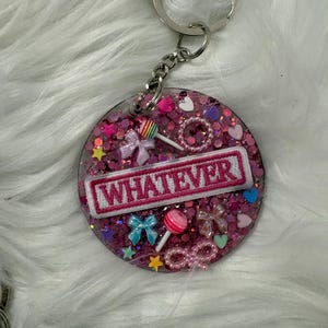May include: A circular keychain featuring a pink glitter-filled resin base with a white embroidered patch that reads WHATEVER in pink letters. The surface is decorated with small charms including bows, lollipops, hearts, stars, and beaded loops.