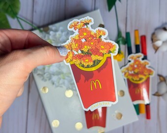 Pode incluir: Um autocolante com um recipiente vermelho de batatas fritas McDonald's cheio de flores laranja e amarelas. O logótipo McDonald's é visível, e o texto "food is fuel" está impresso no recipiente. Outros autocolantes e canetas estão no fundo.