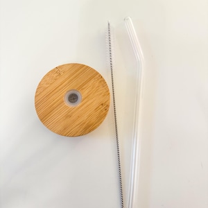 Puede incluir: Una tapa de bamb&uacute;, una pajita de cristal transparente y un cepillo de limpieza de pajitas est&aacute;n dispuestos sobre una superficie blanca. La tapa es redonda con un peque&ntilde;o agujero en el centro. La pajita es curva. El cepillo es plateado.