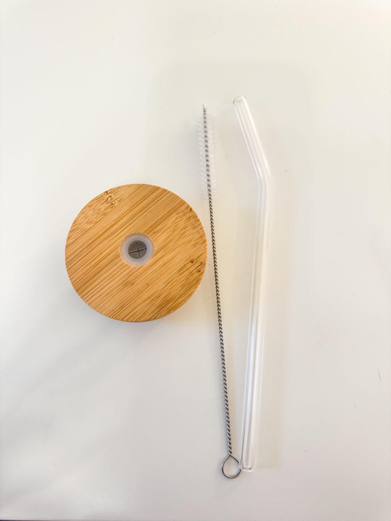 Puede incluir: Una tapa de bamb&uacute;, una pajita de cristal transparente y un cepillo de limpieza de pajitas est&aacute;n dispuestos sobre una superficie blanca. La tapa es redonda con un peque&ntilde;o agujero en el centro. La pajita es curva. El cepillo es plateado.