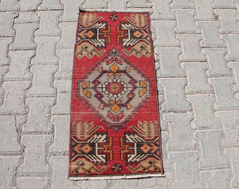 May include: A red, orange, and black patterned rug with a geometric design. The rug has a fringe border and is lying on a gray concrete surface.