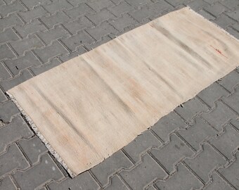 Puede incluir: Una alfombra tejida lisa de color blanco roto con flecos en los bordes. La alfombra está tendida sobre una superficie de hormigón gris.