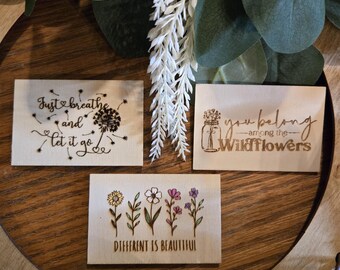 Op de afbeelding: Drie houten platen met inspirerende citaten en bloemmotieven. Eén plaat leest "Just breathe and let it go" met een paardenbloem. Een andere zegt "you belong among the wildflowers" met een pot met bloemen. De derde zegt "DIFFERENT IS BEAUTIFUL" met bloemenillustraties.