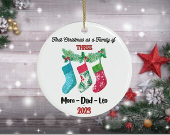 May include: White ceramic Christmas ornament with a red, green, and white stocking design. The ornament reads "First Christmas as a Family of Three" and "Mom - Dad - Leo 2023".