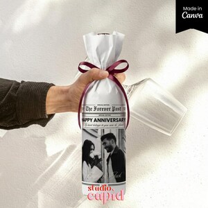 May include: A wine bottle wrapped in white paper with a dark red ribbon, featuring a black and white photo of a couple and the text The Forever Post, Happy Anniversary, My heart belongs to you now and forever, and the names Emily and Jack.