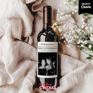 May include: A dark wine bottle with a custom label that reads "EMILY IS TURNING 28!" with a black and white photo. The bottle is set against a backdrop of white fabric and white flowers. The words "studio. cupid" are at the bottom.