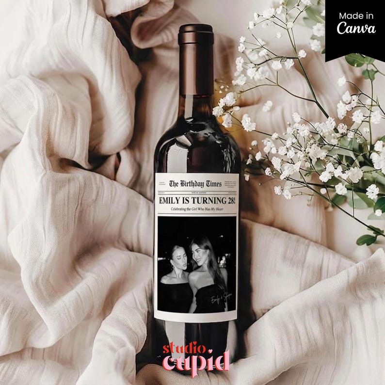 May include: A dark wine bottle with a custom label that reads "EMILY IS TURNING 28!" with a black and white photo. The bottle is set against a backdrop of white fabric and white flowers. The words "studio. cupid" are at the bottom.