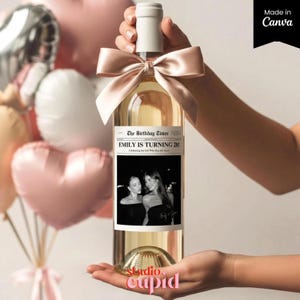 Puede incluir: Una botella de vino transparente con tapón blanco y lazo color crema. La etiqueta dice "The Birthday Times" y "Emily is Turning 28!" con una foto en blanco y negro. La botella se sostiene frente a globos rosas.
