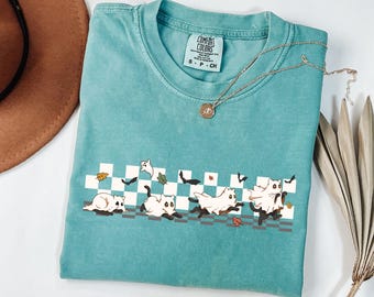 May include: Teal Comfort Colors t-shirt featuring a Halloween-themed design. The graphic shows white cats dressed as ghosts running across a checkered pattern with bats and leaves. A gold necklace with a pendant is draped across the shirt.