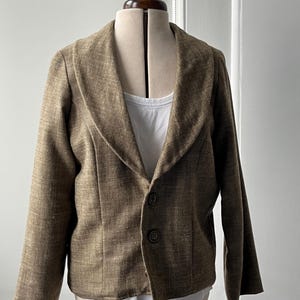 May include: A brown blazer with a notched lapel and a two-button fastening. The jacket is displayed on a mannequin, revealing a white top beneath. The fabric has a textured appearance.