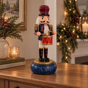 May include: A wooden nutcracker figurine standing on a round blue base decorated with gold stars. The nutcracker wears a red hat, a blue jacket with gold fringe, and black boots, while holding two drumsticks over a red and gold drum.
