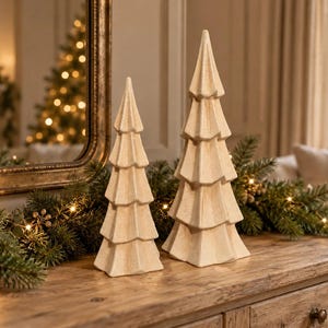May include: Two cream-coloured ceramic Christmas tree figurines with a textured, speckled finish. The trees feature a tiered, geometric design with pointed tops and a wide base. They are positioned side-by-side, with one tree slightly taller than the other.