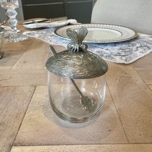 Op de afbeelding: Een zilveren honingpot met een bijenontwerp op de deksel en een klein lepeltje erin. De pot is van glas en heeft een helder, getextureerd oppervlak.
