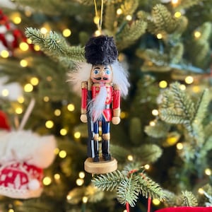 May include: A festive Christmas tree decoration featuring a nutcracker soldier. The ornament has a red jacket, blue trousers, and black boots. It has a black hat, white hair, and a moustache. The decoration hangs from a gold string.
