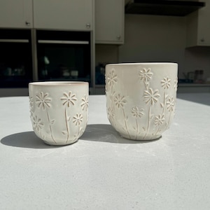 May include: Two off-white ceramic flower pots. The pots feature an embossed daisy design on a textured surface. The smaller pot is on the left, and the larger pot is on the right. The pots are on a light grey surface.