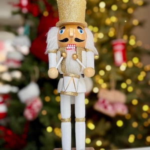 Pode incluir: Figura de quebra-nozes de madeira branca e dourada com chapéu alto dourado brilhante e rosto pintado. O quebra-nozes segura baquetas e está sobre uma base prateada. O fundo é uma árvore de Natal desfocada.