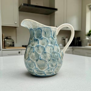 May include: A ceramic pitcher featuring a rounded body and a curved handle. The exterior surface is decorated with a repeating blue and white scale-like pattern, while the interior and handle are finished in a solid, speckled off-white glaze.