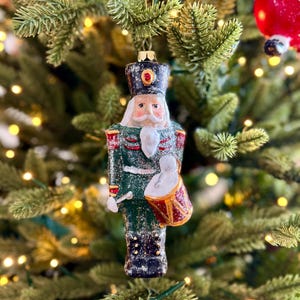 May include: A glass Christmas tree ornament in the shape of a nutcracker soldier. The ornament is green, red, black, and white, with a gold crown and a drum. It hangs on a green Christmas tree with yellow fairy lights.