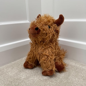 May include: A plush Highland cow toy featuring long, shaggy, ginger-brown fur, a dark brown snout, small pointed ears, and black button eyes. The stuffed animal is depicted in a seated position with a textured, curly coat and soft limbs.