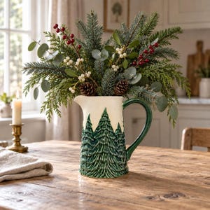 Peut inclure: Un pichet en céramique avec une partie supérieure crème et un motif en relief de sapins vert foncé autour de la base. Le pichet est rempli d'un arrangement festif de branches de sapin, feuilles d'eucalyptus, baies rouges, baies blanches et pommes de pin.
