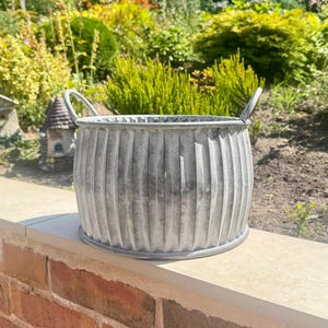May include: A round, silver-coloured metal planter with a vertical ribbed texture and two small handles on opposite sides. The container has a weathered, galvanised finish and a wide, open top, designed for holding plants or decorative items.