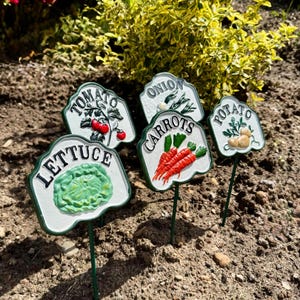 Puede incluir: Un conjunto de marcadores decorativos para jardín con fondos blancos y bordes verdes. Cada marcador presenta una ilustración de una verdura diferente: lechuga, tomate, cebolla, zanahorias y patata. Los marcadores están colocados en el suelo.