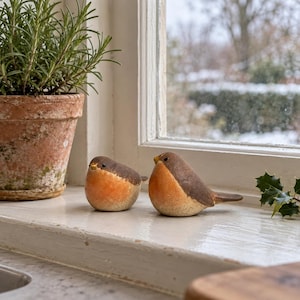 May include: Two small, rounded bird figurines with brown heads and backs, orange-toned chests, and light-colored bellies. The birds have small, dark eyes and pointed beaks, positioned side-by-side on a white surface near a terracotta plant pot.