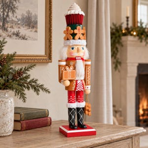 May include: A decorative nutcracker figure featuring a cupcake-shaped hat topped with white frosting, adorned with gingerbread man accents. The figure wears a brown jacket with white icing details, holding a gift box, and black boots.