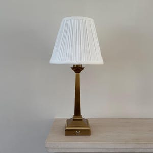 May include: A table lamp featuring a tall, slender, brushed brass-toned metal base with a square, stepped pedestal and a white, pleated, conical fabric lampshade. The base includes a small, circular power button on the front of the square bottom.