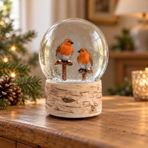Könnte beinhalten: Eine Schneekugel aus Glas mit zwei orangefarbenen und grauen Rotkehlchen, die auf kleinen Holzpfosten sitzen. Der Sockel der Kugel ist wie ein Stück strukturiertes weißes Birkenholz gestaltet und das Innere enthält weiße Schneepartikel.
